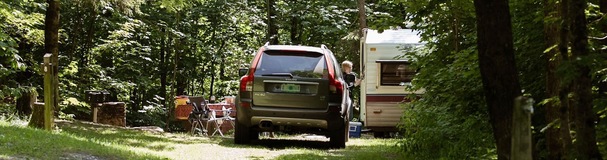 The image shows a forested campsite with a parked dark green Volvo XC90 SUV and a white camper trailer. A person with short blonde hair is standing by the camper's door. Nearby, there is a picnic table with a red tablecloth, folding chairs, and camping supplies. A metal grill and a numbered campsite marker ("8") are also visible. The area is lush with green trees, and sunlight filters through the foliage, creating a peaceful outdoor setting.