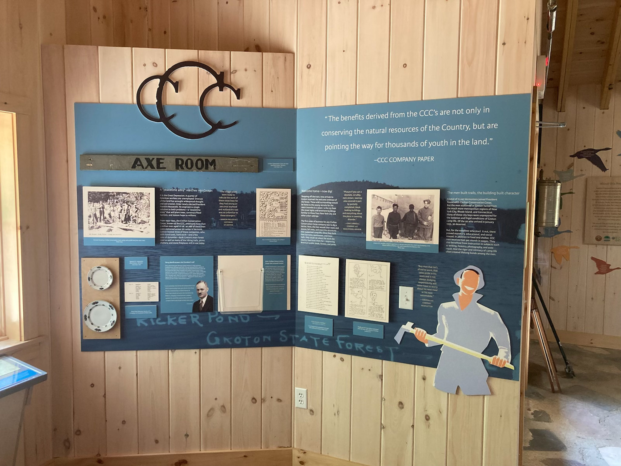 The image shows an informational display about the Civilian Conservation Corps (CCC) inside a wooden-paneled room. The display features a large blue panel with the letters "CCC" prominently displayed at the top. Below, there is a wooden sign labeled "AXE ROOM." The panel includes historical photographs, documents, and text describing the CCC's contributions to conservation and youth development. 