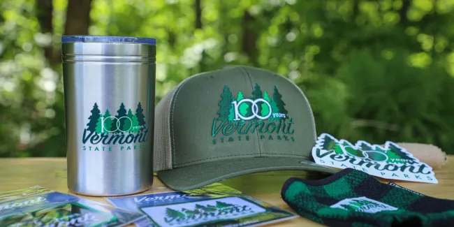 A display of 100th anniversary branded merchandise including stickers, a hat, a mug, and a koozie.