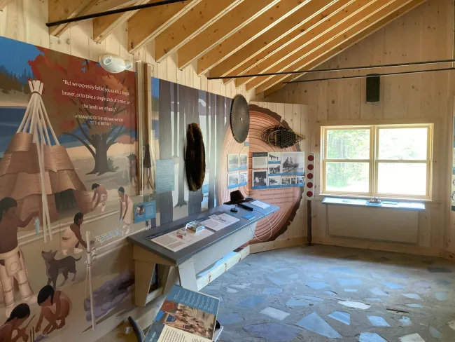 The image depicts an indoor exhibit at a nature center or museum. The walls are adorned with murals and informational panels. On the left, a mural illustrates a scene with Indigenous people, traditional structures, and a dog, accompanied by a quote from an Abenaki Nation statement to the British, emphasizing the protection of natural resources. The center of the exhibit features a display case with artifacts and educational materials.