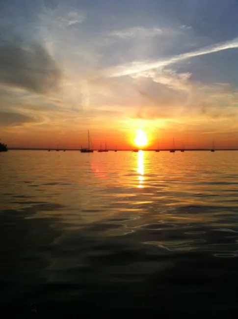 A breathtaking sunset over a calm body of water, with the sun casting a warm golden glow across the sky and reflecting on the water's surface. Several sailboats are silhouetted against the horizon, adding a serene and picturesque element to the scene. Wispy clouds are scattered across the sky, catching the colors of the sunset and enhancing the tranquil atmosphere.