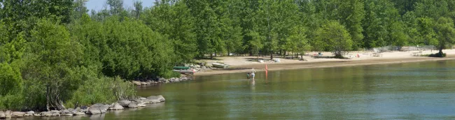 The image shows a calm lakeside beach bordered by green trees. In the foreground, shallow water reflects the surrounding foliage. A man and child stand by the water’s edge, with a small boat pulled onto the sandy shore nearby. Picnic tables and a few beach chairs are scattered in the background, with an orange cone placed near the water for marking. Rocks and plants line the shore, giving the scene a peaceful, natural feel ideal for relaxation and family time outdoors.
