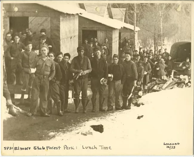 This black-and-white photo from 1933 shows a large group of Civilian Conservation Corps (CCC) workers at Elmore State Forest Park in Vermont, lined up for lunch in winter. The men, dressed in heavy work clothes and boots, carry tin lunch pails and mugs. They stand on a snow-lined dirt road beside rustic wooden buildings, with chopped logs piled nearby. The handwritten caption reads, "SP3/Elmore State Forest Park: Lunch Time."